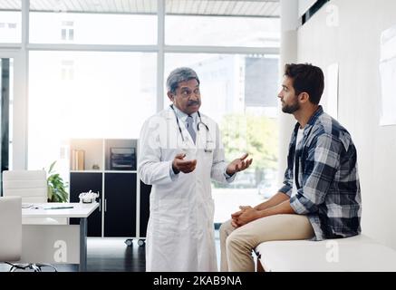 Pouvez-vous m'en dire plus sur vos symptômes.un médecin et un patient hommes matures ayant une discussion au bureau des médecins avant un examen. Banque D'Images