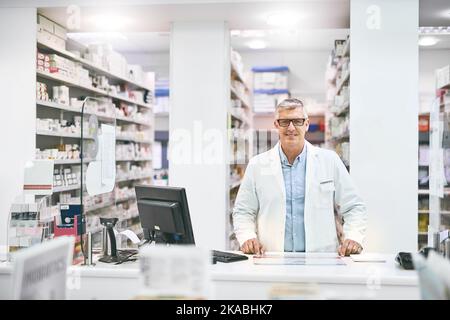 Nous avons des quantités infinies de médicaments. Portrait d'un pharmacien homme mature et gai debout derrière le comptoir tout en regardant la caméra dans un Banque D'Images