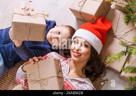 Joyeux maman avec son fils au chapeau de père noël se trouve sur le sol à la maison sous l'arbre avec des cadeaux, profitez de noël et de vacances du nouvel an. Une famille joyeuse la veille de noël Banque D'Images