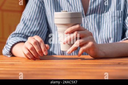 Femme porte une tasse de café écologique à emporter en silicone réutilisable dans les mains au-dessus d'une table en bois. Banque D'Images