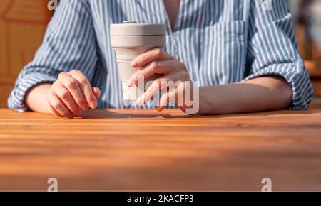 Femme porte une tasse à café écologique réutilisable en silicone à emporter dans les mains au-dessus d'une table en bois. Banque D'Images