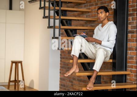 Jeune homme en vêtements blancs assis sur des escaliers en bois et prenant des notes Banque D'Images