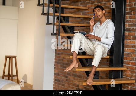 Jeune homme en vêtements blancs assis sur des escaliers en bois et prenant des notes Banque D'Images