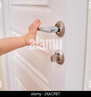 Tout-petit ouvre la porte maintenant la poignée de porte, la main de l'enfant en gros plan. Porte en bois blanc, poignée de porte en métal et main de bébé Banque D'Images