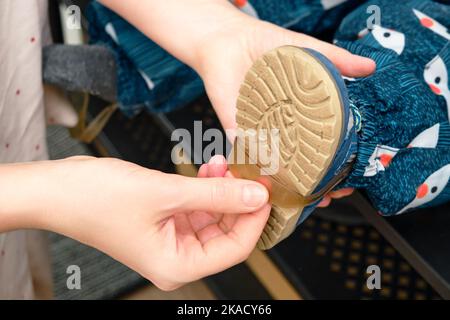 La mère met une bande pour les chaussures sur le pied bébé tout-petit assis dans le couloir de la maison. Femme maman vêtue chaud bottes vêtements sur enfant pour la marche d'hiver Banque D'Images