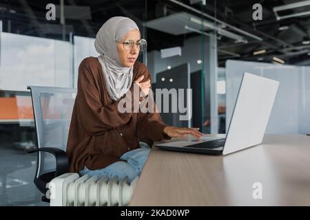 Femme d'affaires qui gèle au bureau au travail, femme dans le hijab près du chauffage essayant de rester au chaud travaillant à l'intérieur du bâtiment de bureau avec ordinateur portable, froid dans le bureau. Banque D'Images