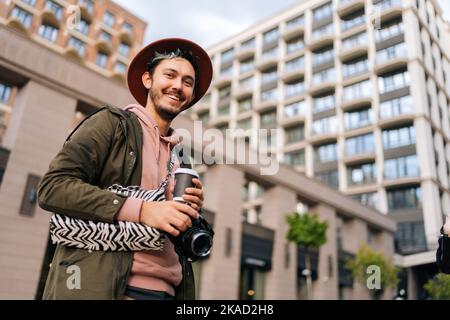 Vue en contre-angle d'un jeune homme joyeux barbu portant une femme élégante chapeau tenant dans l'appareil photo à main, tasse à café et sac à main de dame, souriant regardant l'appareil photo. Banque D'Images