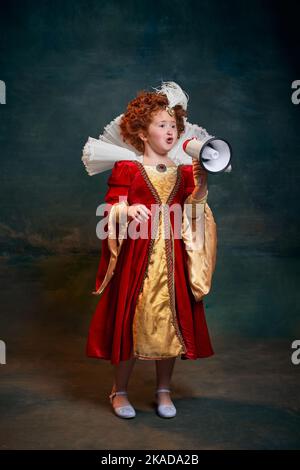 Portrait de petite fille à tête rouge, enfant en costume de personne royale parlant en mégaphone isolé sur fond vert foncé Banque D'Images