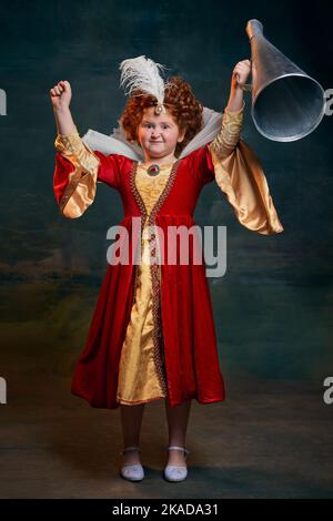 Portrait de petite fille à tête rouge, enfant en costume de personne royale isolé sur fond vert foncé. Look gagnant Banque D'Images