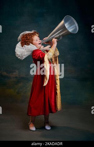 Portrait d'une petite fille à tête rouge, enfant en costume de personne royale criant en mégaphone isolé sur fond vert foncé Banque D'Images