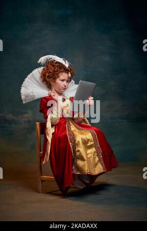 Portrait d'une petite fille à tête rouge, enfant en costume de la personne royale dactylographiant sur une tablette isolée sur fond vert foncé Banque D'Images