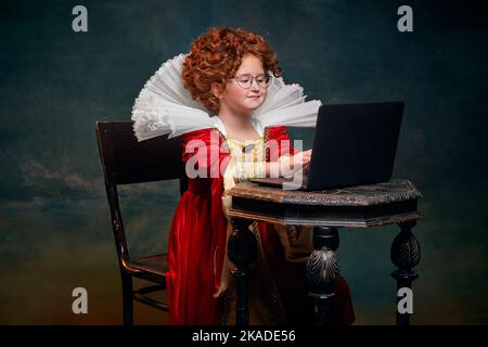 Portrait d'une petite fille à tête rouge, enfant en costume de la personne royale dactylographiant sur ordinateur portable isolé sur fond vert foncé Banque D'Images