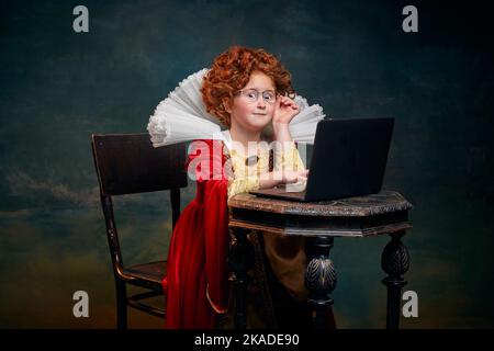 Portrait d'une petite fille à tête rouge à l'image d'une personne royale avec ordinateur portable isolé sur fond vert foncé. Étudier Banque D'Images