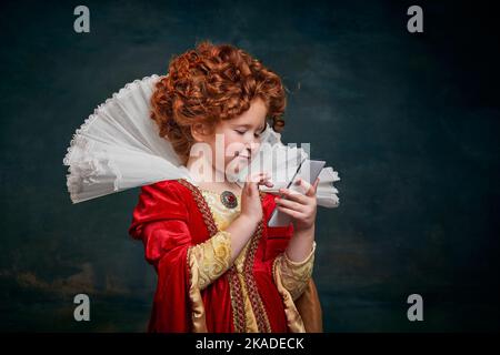 Portrait de petite fille à tête rouge, enfant en costume de personne royale avec téléphone isolé sur fond vert foncé Banque D'Images