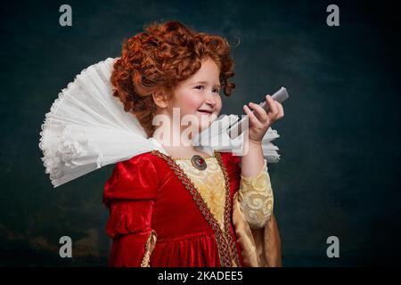 Portrait d'une petite fille à tête rouge, personne royale enregistrant un message vocal sur un téléphone isolé sur fond vert foncé Banque D'Images