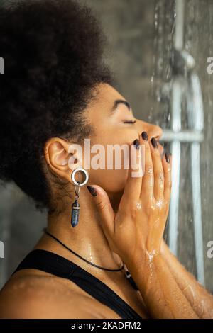 Vue latérale de femme afro-américaine lavant le visage sous l'eau dans la douche, image de stock Banque D'Images