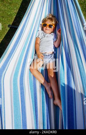 Petit garçon blond souriant avec lunettes de soleil, repose dans un hamac Banque D'Images