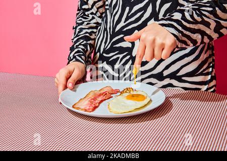 Femme mettant le doigt à l'intérieur du jaune d'œuf. Assiette de petit déjeuner anglais sur nappe rose Banque D'Images