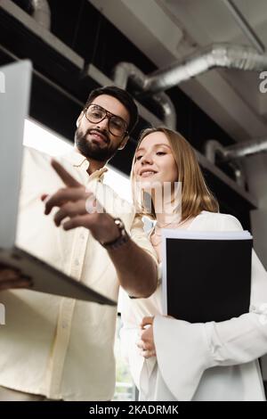 Vue à angle bas d'un homme d'affaires pointant vers un ordinateur portable près d'un nouvel employé avec dossier en papier dans le bureau, image de stock Banque D'Images