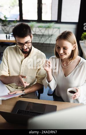 Un nouvel employé tenant une tasse de café près d'un homme d'affaires avec des documents pointant vers un ordinateur portable au bureau, image de stock Banque D'Images