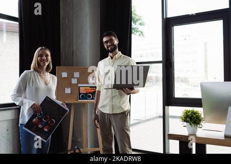 Manager souriant tenant un ordinateur portable et regardant la caméra près d'un interne avec des cartes dans le bureau, image de stock Banque D'Images