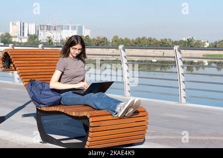 Une adolescente est assise sur un banc de parc avec un ordinateur portable et se prépare pour des leçons ou des examens. Une fille étudiante espagnole avec des lunettes tapant du texte sur un lapto Banque D'Images