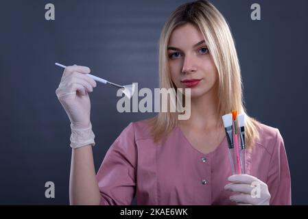 une jeune fille médecin tient des brosses cosmétiques pour les procédures de peau dans ses mains. Sur fond gris Banque D'Images