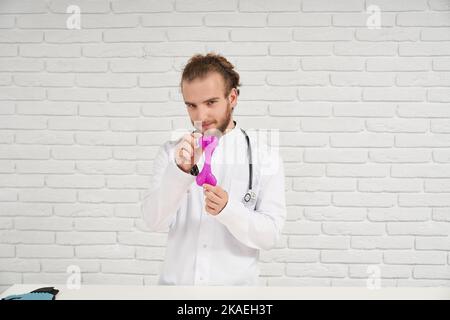 Portrait de vétérinaire masculin en robe blanche avec cheveux bouclés dans le bandeau montrant le jouet de chien rose en caoutchouc pour mâcher pendant la dentition. Véritable spécialiste toujours prêt à aider nos petits frères - animaux. Banque D'Images