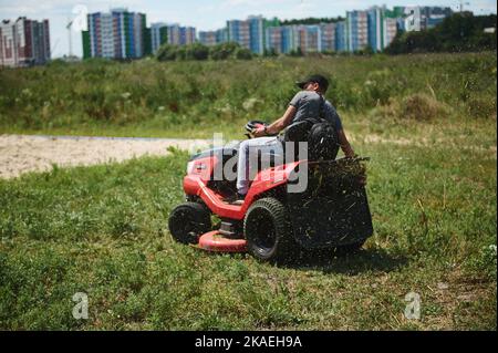 Un jardinier sur une tondeuse à gazon, un équipement électrique pour tondre l'herbe verte, entretenir les parcs, contre les bâtiments urbains élevés Banque D'Images