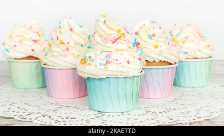 Cupcakes à la vanille et crème au beurre garnies de saupoudrées d'or comestibles en gros plan. Les muffins dans des gobelets en papier coloré se tiennent sur une serviette en dentelle blanche sur un fe Banque D'Images