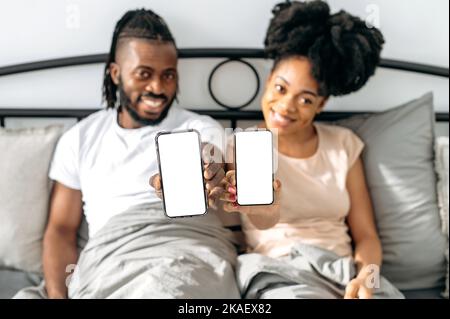 Joyeux couple afro-américain positif, heureux conjoints, s'asseoir dans un lit confortable dans la chambre, montrer leurs smartphones avec des écrans de maquette vierges, espace pour la publicité, la présentation, regarder l'appareil photo, sourire Banque D'Images