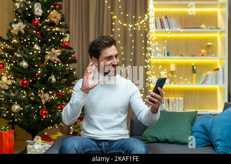 Salutations en ligne avec les vacances du nouvel an. Jeune homme beau à la maison sur le canapé parlant sur un appel vidéo, félicitant pour les vacances de Noël. Tient le téléphone dans ses mains, fait un bruit de main. Banque D'Images