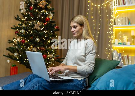 Célébration en ligne. Une jeune belle femme célèbre le nouvel an, Noël en ligne, à distance avec sa famille et ses amis par appel vidéo depuis un ordinateur portable. Asseyez-vous à la maison sur le canapé près de l'arbre de Noël. Banque D'Images