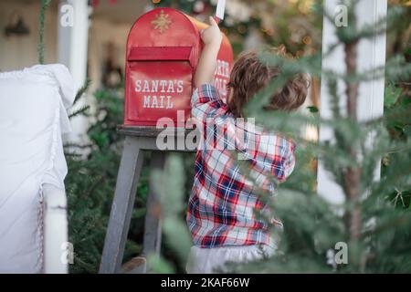 Un tout-petit touchant la boîte aux lettres d'un Père Noël sur une chaise avec un sapin au premier plan Banque D'Images