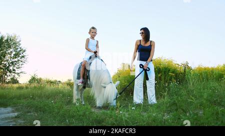 Une femme et un garçon marchent sur le terrain, le fils est à cheval sur un poney, la mère tient un poney pour une bride. Bonnes vacances en famille. À l'extérieur, en été, près de la forêt. Photo de haute qualité Banque D'Images