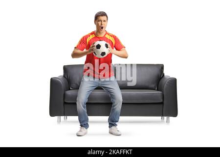 Jeune homme excité devant un canapé noir tenant un ballon de football et applaudisant isolé sur fond blanc Banque D'Images