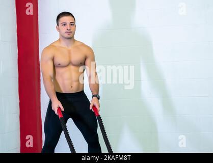 Un beau jeune homme musclé utilisant des cordes de combat pour l'exercice dans une salle de gym. Il se concentre sur la réalisation de l'exercice. Photo de motivation. Copier l'espace. Banque D'Images