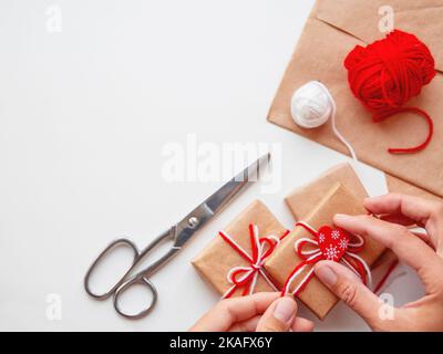 Les cadeaux de bricolage sont emballés dans du papier artisanal. Cadeaux avec fils blancs et rouges et cœur rouge. Boîtes en mains sur fond blanc. Banque D'Images