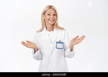 Femme aîée élégante et souriante, médecin invitant les patients à la clinique, à l'air heureux et optimiste, debout sur fond blanc. Banque D'Images