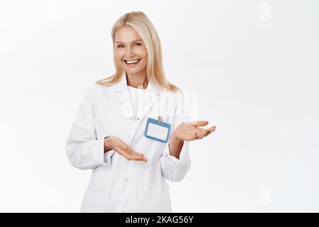 Femme aîée élégante et souriante, médecin invitant les patients à la clinique, à l'air heureux et optimiste, debout sur fond blanc. Banque D'Images