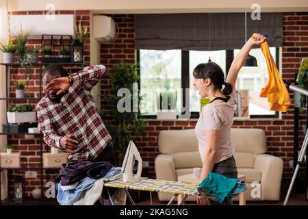 Une femme irritée hurle à son mari à la maison tout en repassant des vêtements dans la salle de séjour. Couple multiracial arguant sur les corvées dans la maison, l'homme ne pas aider avec les corvées et l'entretien de ménage. Banque D'Images