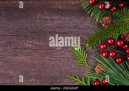 Bordure latérale de Noël avec branches à feuilles persistantes, baies rouges et cônes de pin. Vue en hauteur sur un arrière-plan en bois sombre avec espace de copie. Banque D'Images