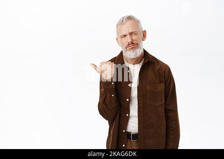 Vieil homme triste et sombre, modèle masculin senior pointant du doigt vers la gauche et grimaquant, se plaignant de mauvaise qualité, debout sur fond blanc Banque D'Images