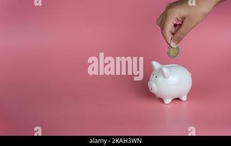 Une main humaine dépose une pièce dans une banque de porc blanche. Sur fond rose. Les concepts d'épargne et d'investissement. Banque D'Images