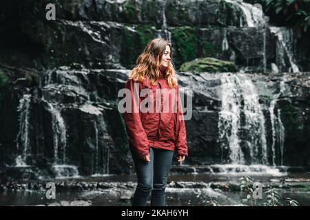 blonde caucasienne fille dans un imperméable rouge et pantalon noir calme et détendu regardant la terre près des chutes d'eau et du lac, purakuauni chutes, nouvelle-zélande Banque D'Images