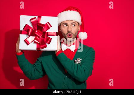 Photo de doux impressionné homme porter x-mas gilet vert devine à l'intérieur noël présent vide espace isolé couleur rouge arrière-plan Banque D'Images