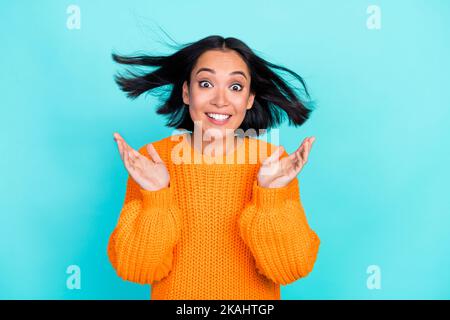 Photo d'adorable fille impressionnée vêtue orange Pullover lever les mains bras air soufflage isolé teal couleur fond Banque D'Images