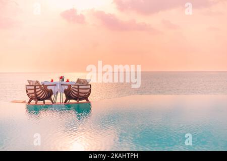 L'été, l'amour et le romantisme table pour un dîner romantique avec des chaises longues à débordement de luxe sous le ciel du coucher du soleil et la mer en arrière-plan Banque D'Images