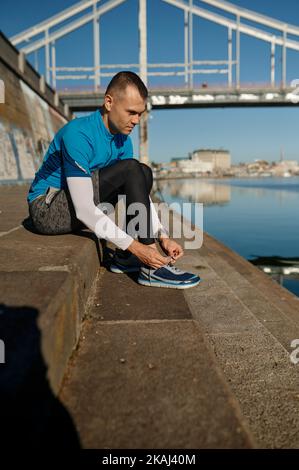 Athlète homme nouant la dentelle assis sur les escaliers avant de s'entraîner sur la rive de la rivière de la ville Banque D'Images
