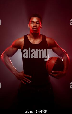 Côté joueur de basket-ball éclairé de couleur rouge tenant une balle sur fond flou. Sérieux homme afro-américain concentré. Banque D'Images